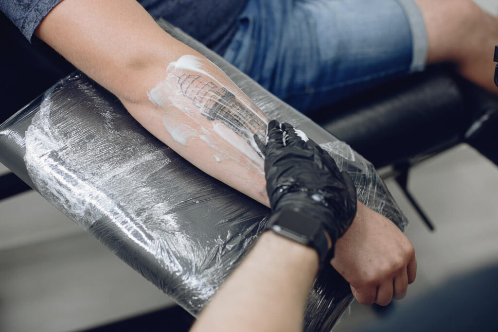 Main appliquant une crème de soin sur un tatouage en cours de cicatrisation, suivant un guide jour après jour.