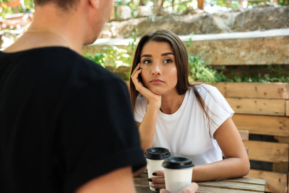 Couple assis à table, l'homme silencieux regarde ailleurs, la femme a l'air ennuyée ou en attente.