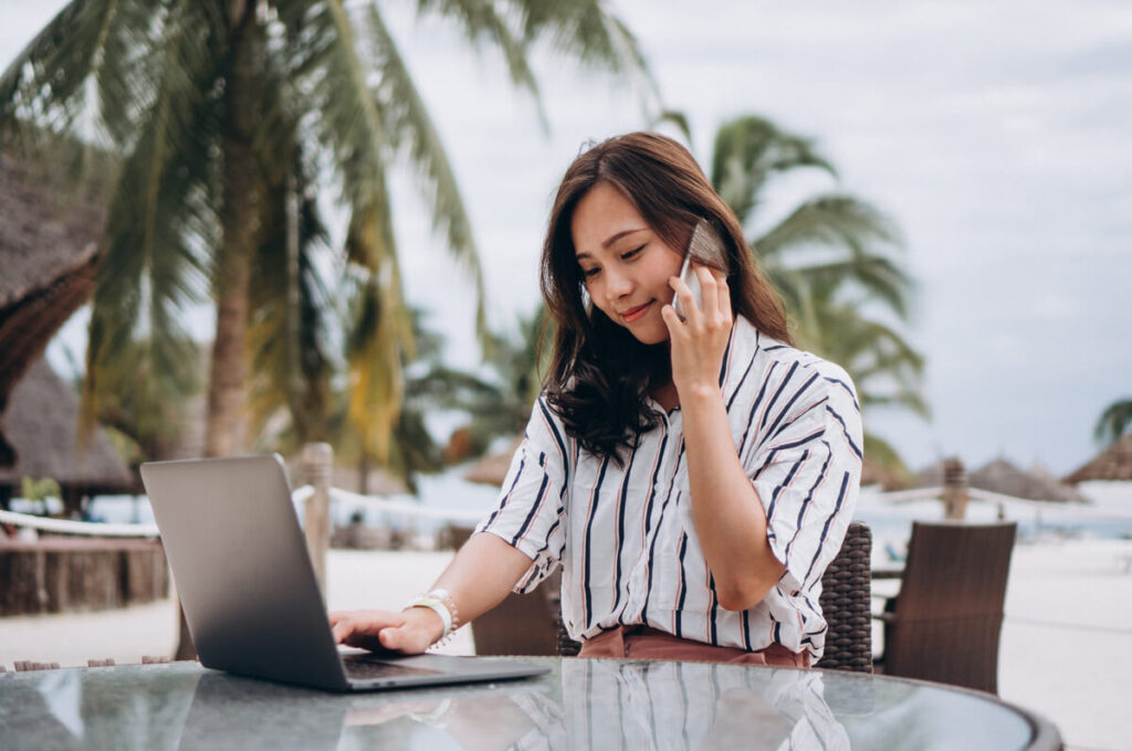 Devenir testeur de voyage : les étapes pour être payé à voyager 1 Une testeuse de voyage travaillant sur son ordinateur portable face à la mer turquoise d'un hôtel de luxe.